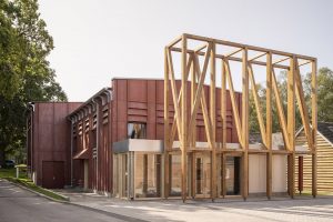 Jonathan Tuckey Design's Horris Hill theatre stands with a low pitched roof and rusty red timber cladding with raised profiles is shown, with a tall timber frame built in front