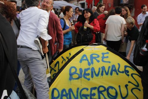 Visitors to the Venice Biennale schmooze around a tent spray-painted with the words 'Are Biennales Dangerous'. Two men actively lean over the tent to air kiss