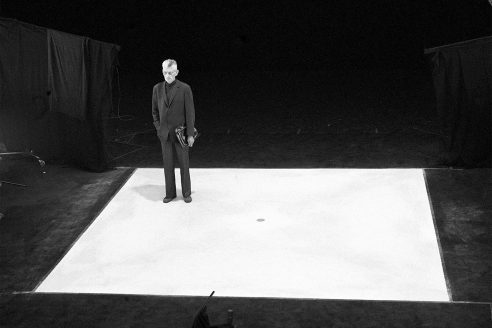 Samuel Beckett stands in the corner of a painted white square, wearing a suit. The surrounding area is all black, so the square and his little white head stand out starkly against the backdrop.