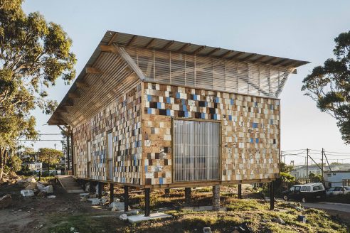 A raised structure is clad with reused, lapped timber tiles, with many painted in different colours