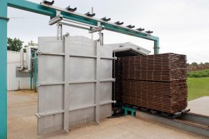 Stacks of lumber are being rolled into a heavy duty steel chamber, its door suspended by a mechanical arm
