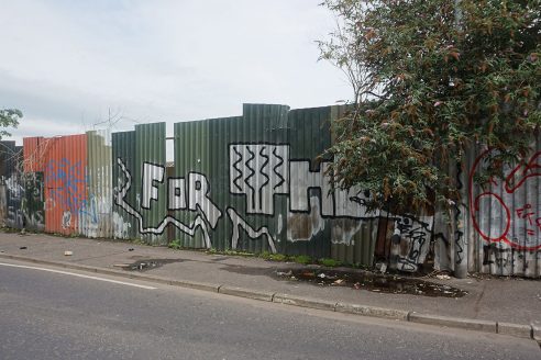 A green corrugated sheet fence is constructed along the side of a pavement in Cupar Way, Belfast