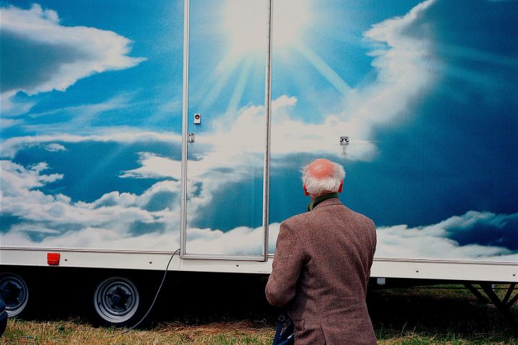 A man appears to kneel on the ground in front of a trailer vehicle plastered with a highly saturated image of the sun over the clouds