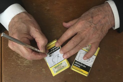 The hands of an elderly man making line drawings of bare feet on two yellow Portuguese cigarette packets