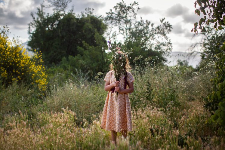 A young girl in a pink dress holds a bunch of flowers while standing in a rich grassland under a clouded sky