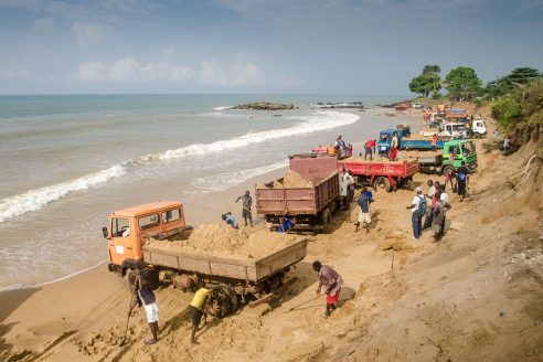 Workers shovel sand into the back of trucks at a beach on Hamilton Beach, Sierra Leone. The sand will be used for concrete, a subject of discussion in Alia Bengana’s new graphic novel