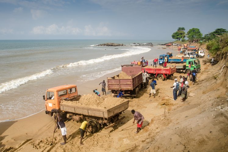 Workers shovel sand into the back of trucks at a beach on Hamilton Beach, Sierra Leone. The sand will be used for concrete, a subject of discussion in Alia Bengana’s new graphic novel