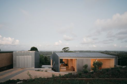 Atelier Chen Hung’s Mapleton House in Australia. Seen from the road at dusk with shutters open, letting out light from inside and showing the deep blue sky and the view to the valley below