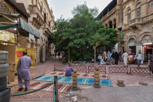 A man is sat on a rug outside a storefront, he is looking towards the row of trees which divide the street. People are walking down the paved road with shops on either side in the ground floor of the buildings, open to the street.
