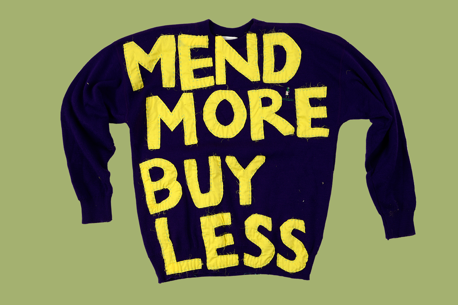 A black jumper on a green background. The jumper has been appliquéd in yellow fabric reading "MEND MORE BUY LESS"